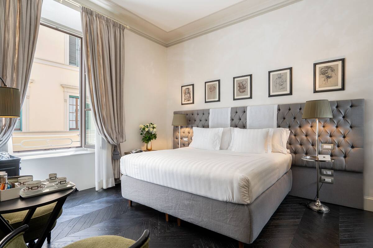 A neatly arranged bedroom with a large bed, a bedside table with a lamp, and a window with curtains.