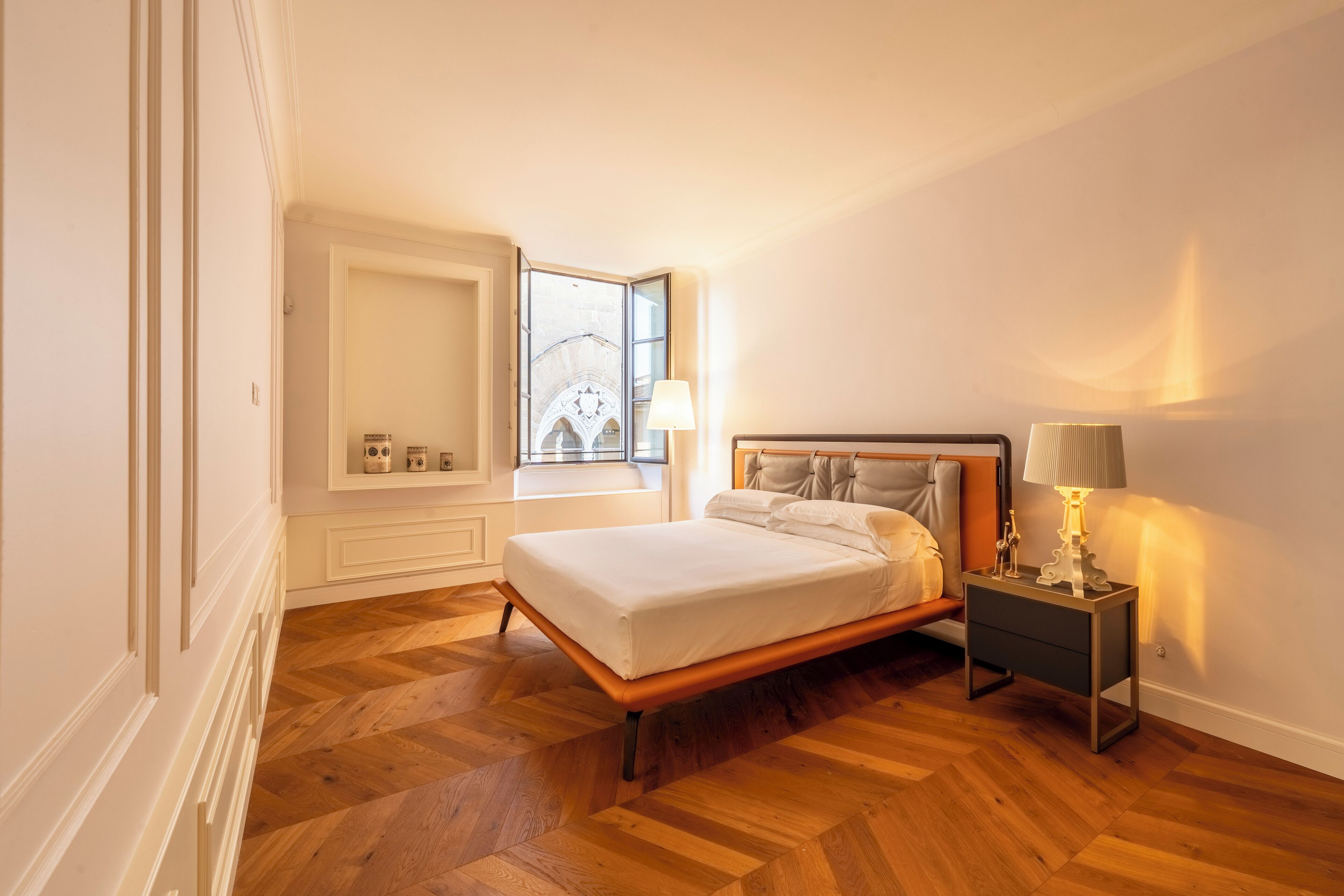 A bedroom with a wooden floor, a bed with white linens, a nightstand with a lamp, and a window with a view of a building.