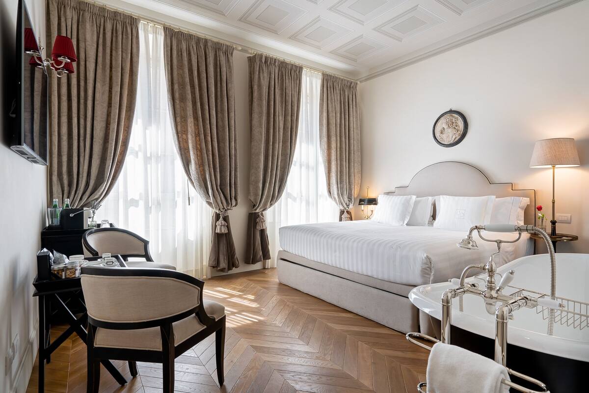 A hotel room with a bed, a desk with a chair, a desk lamp, a bedside table with a lamp, a large window with curtains, and a bathtub.