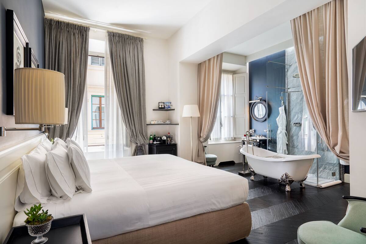 A hotel room with a large bed, a bathtub, a vanity with a mirror, and a window with curtains.