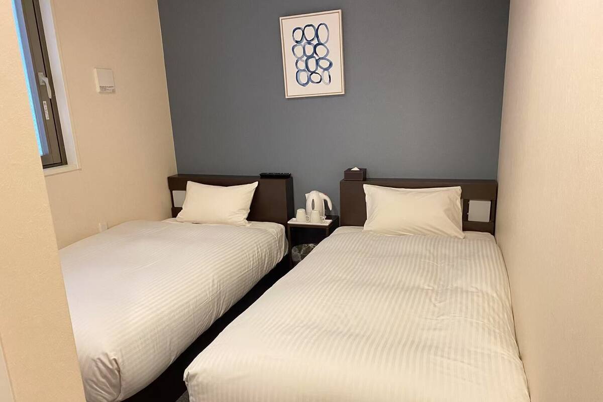 Une chambre d’hôtel avec deux lits simples, une table de chevet avec une lampe, une œuvre d’art encadrée au mur et une fenêtre donnant sur un paysage.