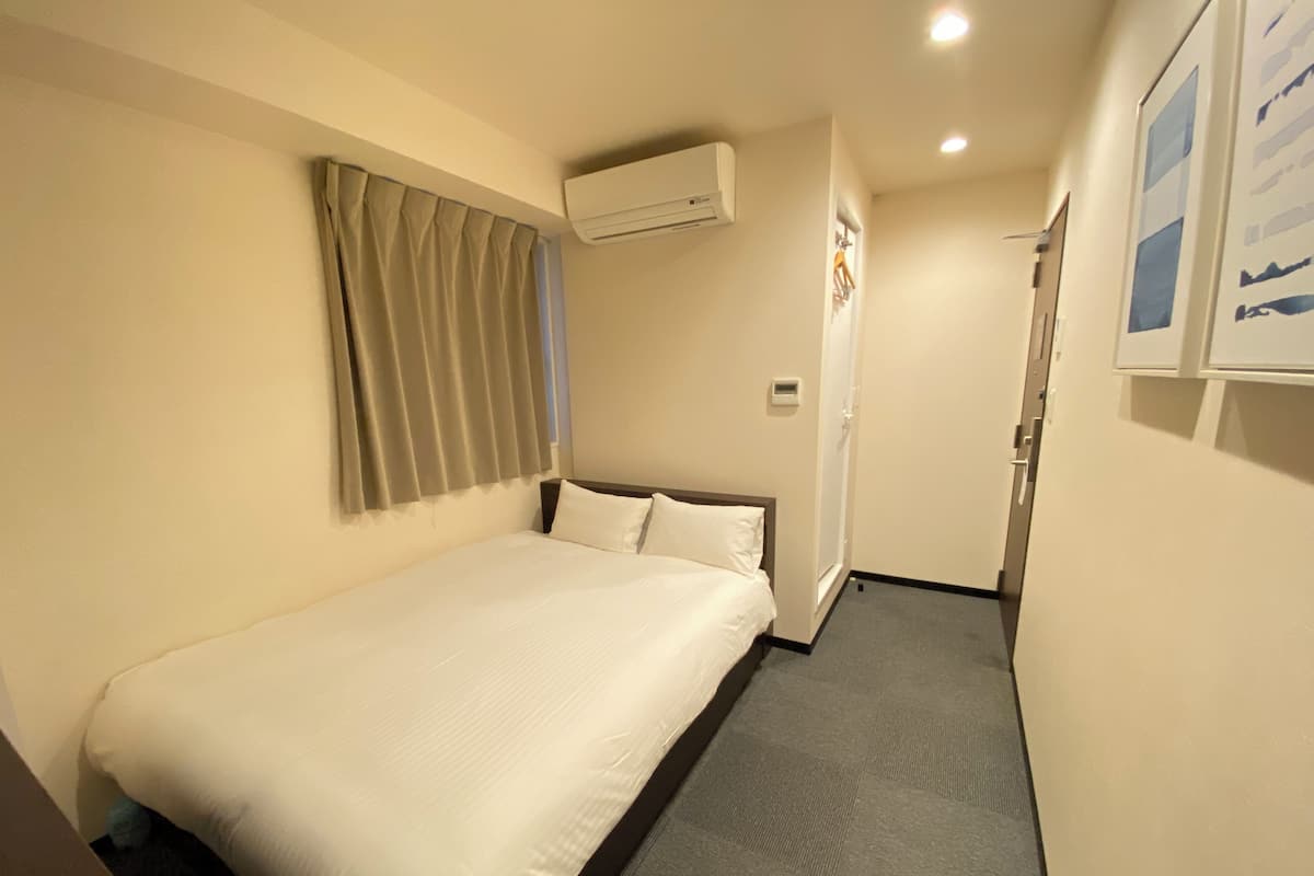 Une chambre d’hôtel avec un lit, un climatiseur fixé au mur, une porte et une petite fenêtre avec des rideaux.