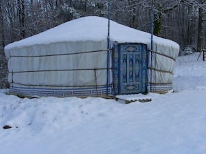 Une yourte enneigée, dotée d’une porte bleue décorative, nichée au cœur d’une forêt couverte de neige.