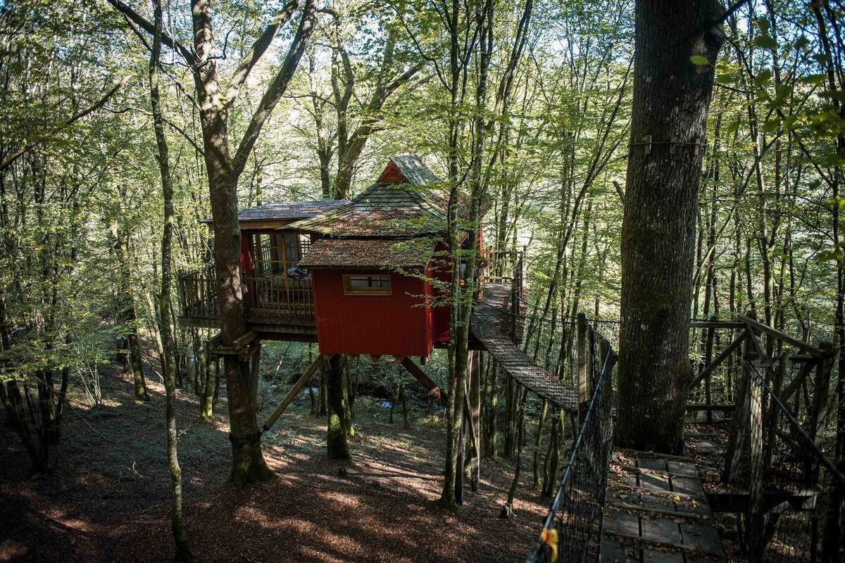 A treehouse with a red exterior, wooden deck, and a small porch area, surrounded by a forested environment.
