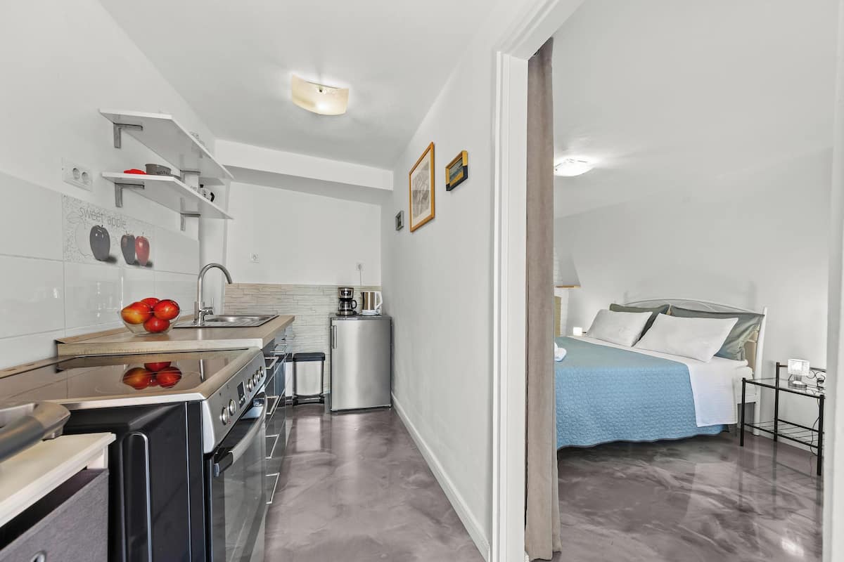 A compact kitchen with modern appliances, a small refrigerator, and a sink. There is a bed with a blue blanket and white pillows in the adjacent room.