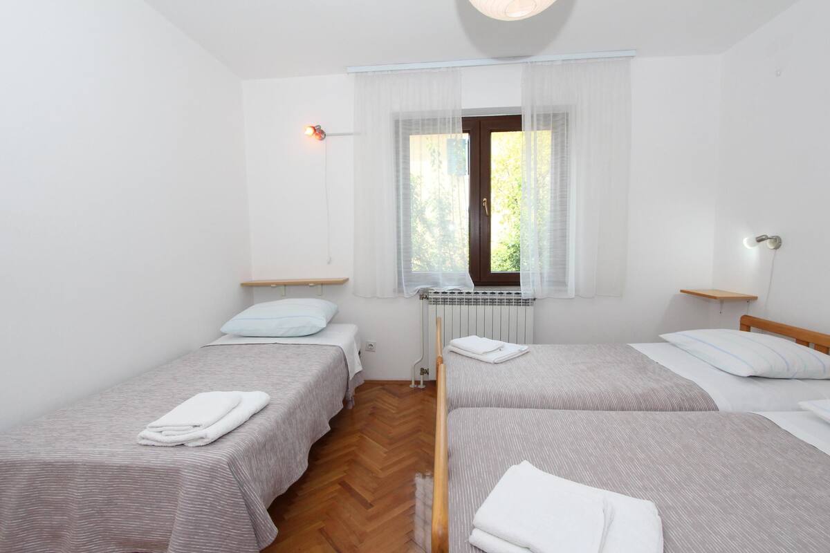 A room with two single beds, a window with sheer curtains, a radiator, and a wall-mounted light fixture.