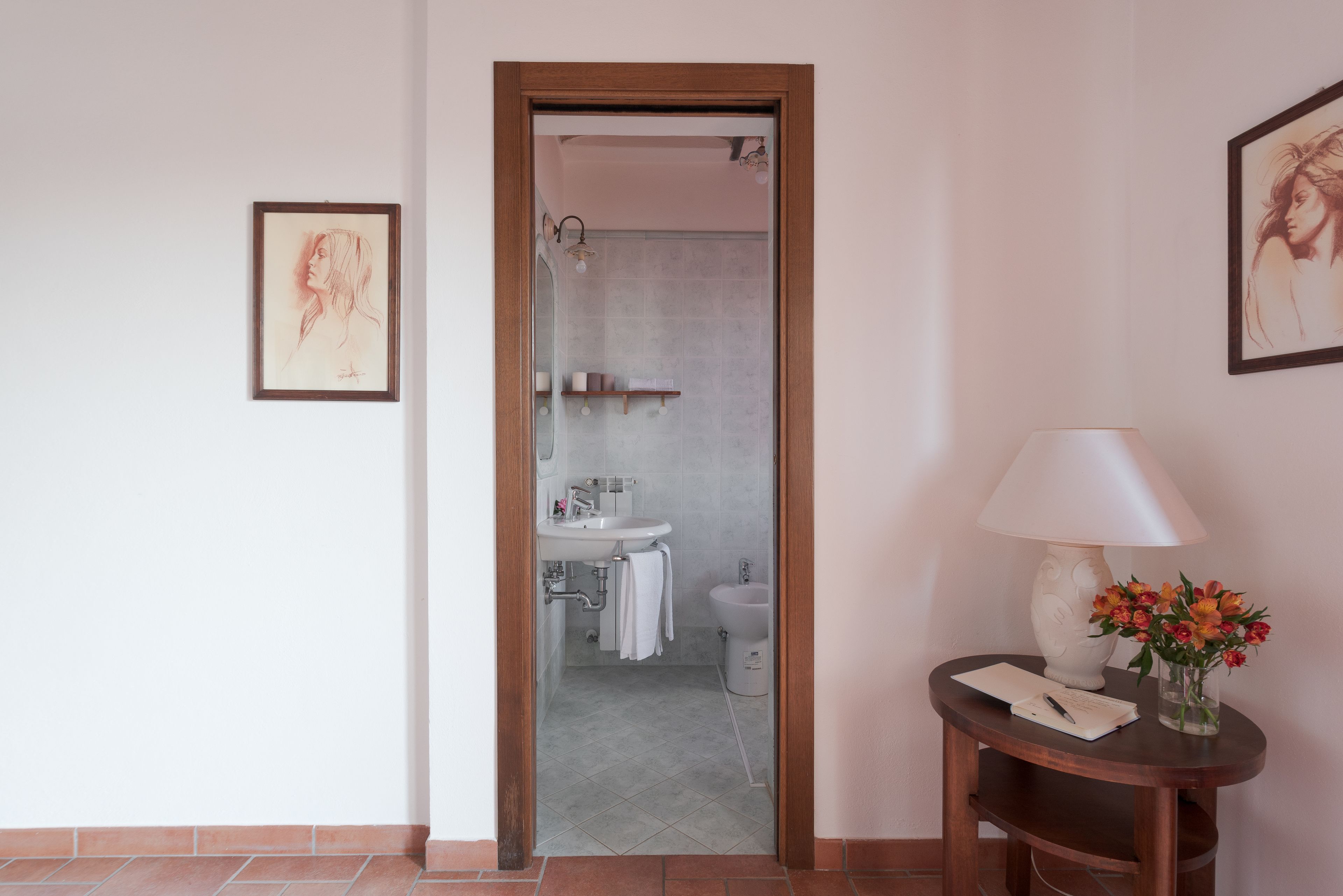 Un baño con lavamanos, inodoro y ducha. Se observa un cuadro en la pared y una mesa con una lámpara y flores.