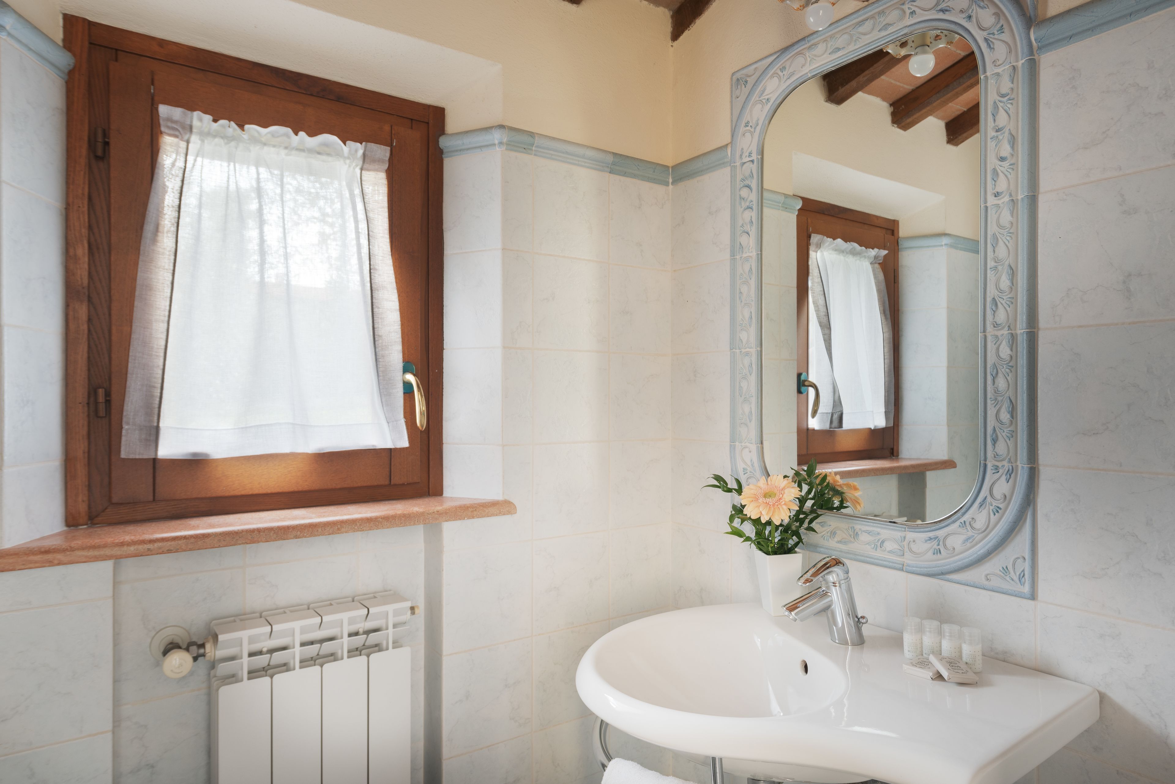 A bathroom with a white sink, a mirror with a decorative frame, a window with a wooden frame, and a radiator.