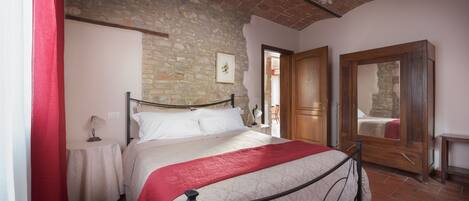 Un dormitorio con pared de piedra, una cama con manta roja, una puerta de madera y una mesita con lámpara.