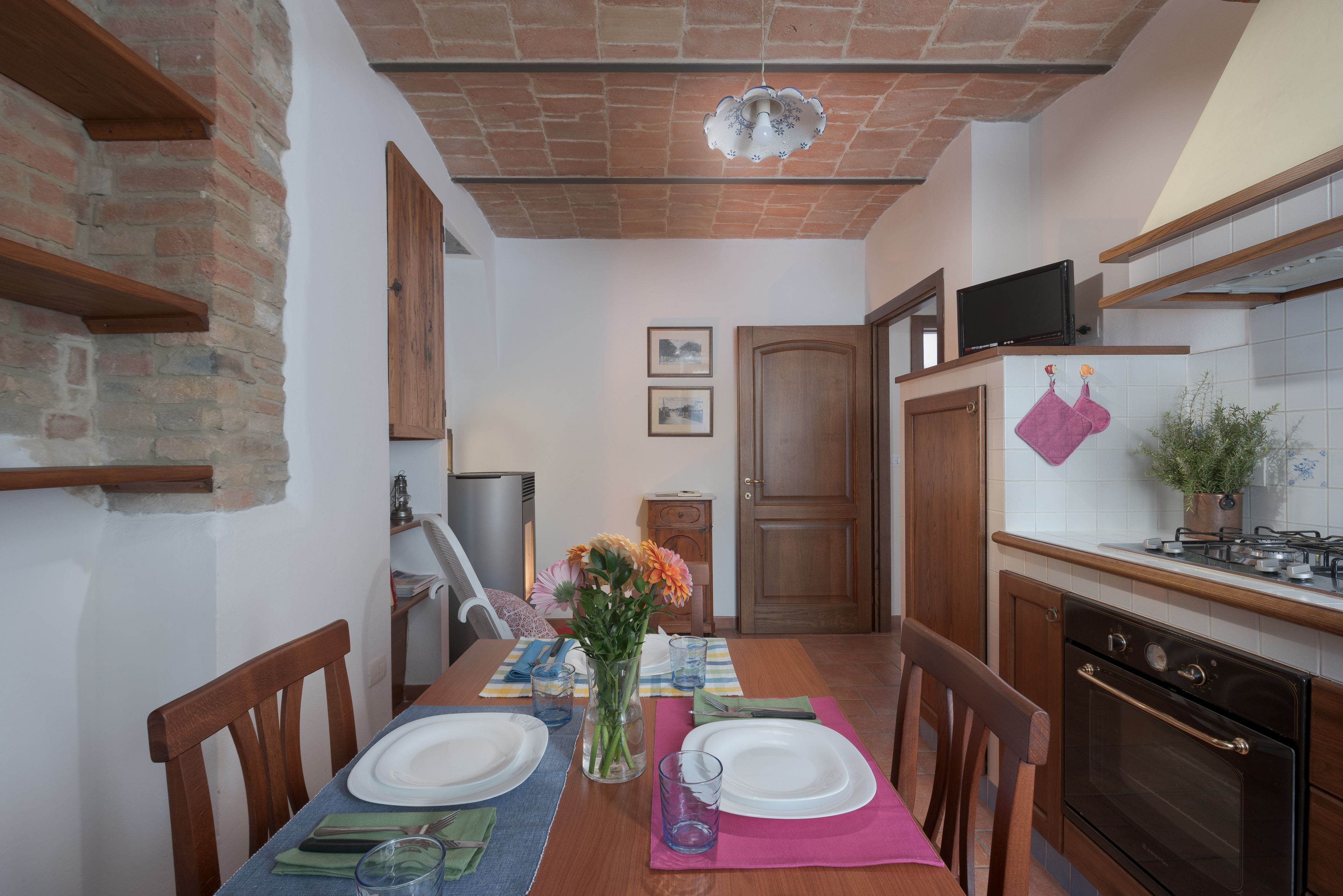 A dining area with a brick wall, wooden chairs, a table set with plates, glasses, and a vase of flowers.