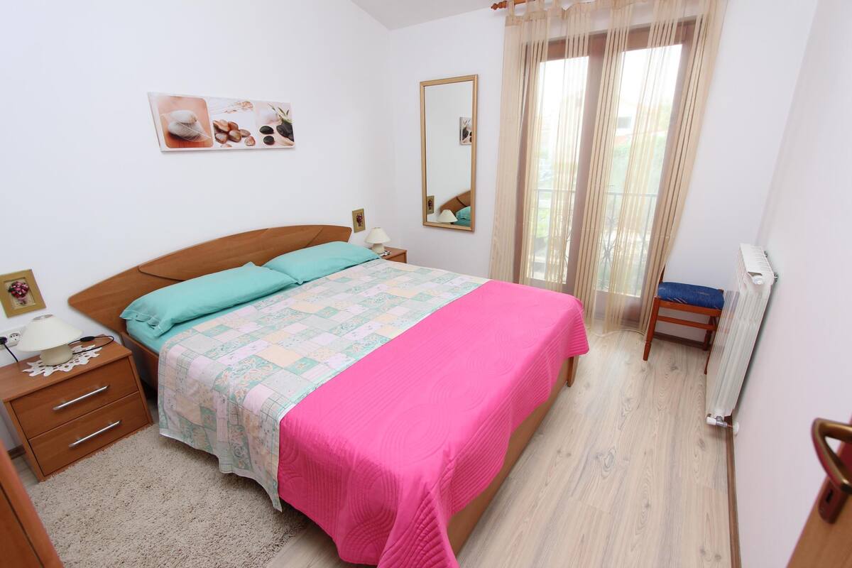 Een slaapkamer met een bed, nachtkastje, stoel, raam met gordijnen en een radiator.