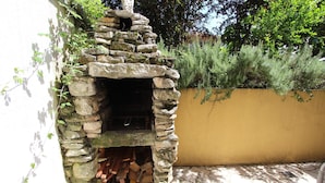 Une cheminée extérieure en pierre, avec du bois de chauffage rangé à l’intérieur, entourée d’un mur jaune et de verdure.