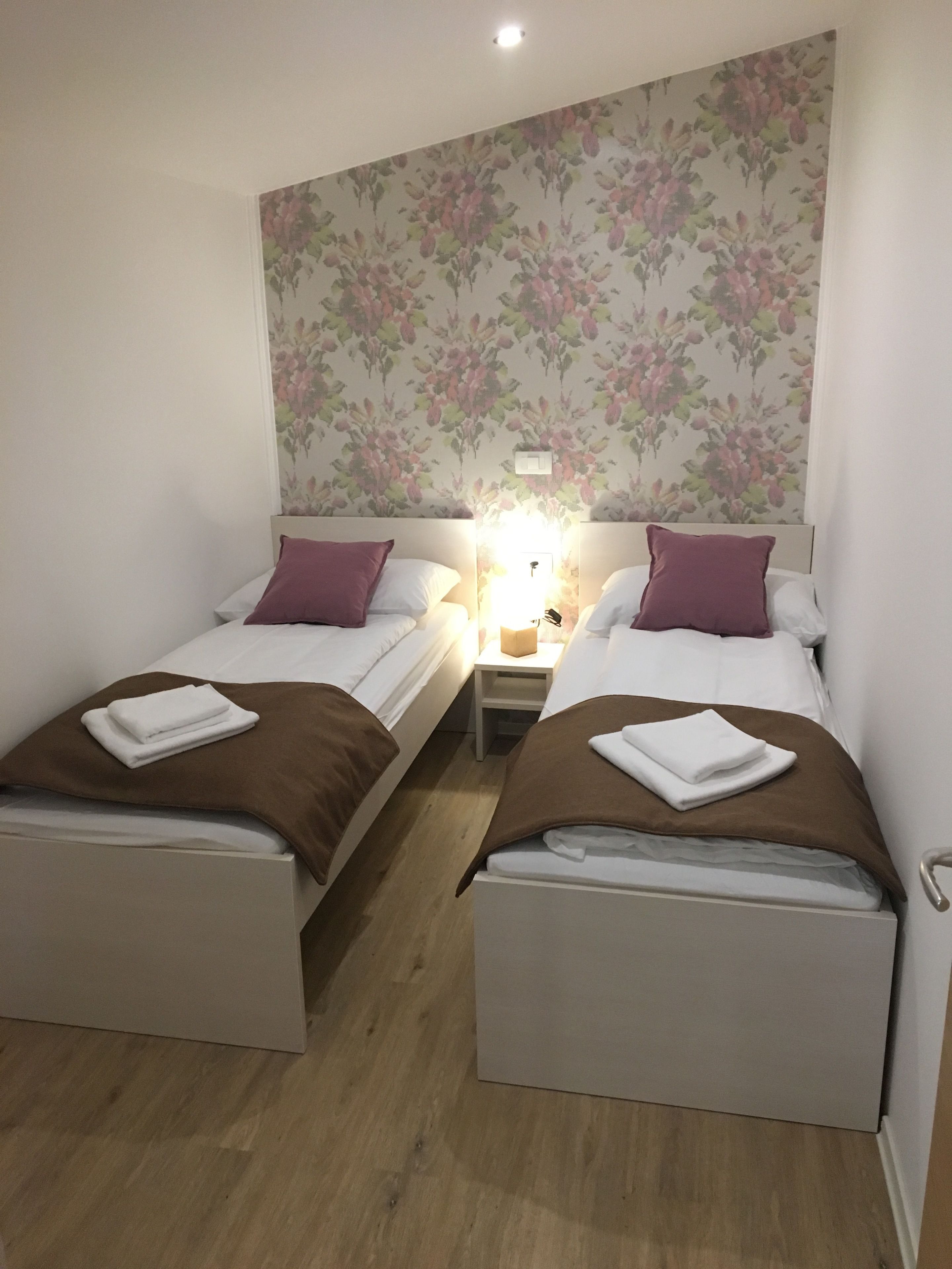 Habitación doble con dos camas individuales, cada una con una manta marrón y almohadas blancas. Entre las camas hay una mesita de noche con una lámpara. La habitación tiene papel tapiz con motivos florales y suelo de madera.