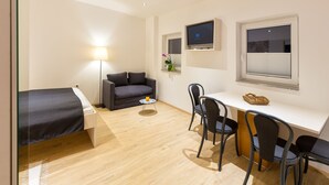 A compact room with a bed, a sofa, a desk with chairs, a TV, and a window with blinds. - Apartments SHISHKA (Ljubljana)