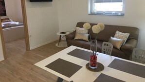 A modern living room with a brown sofa, a dining table with placemats, a TV mounted on the wall, and a window with blinds. - Apartments SHISHKA (Ljubljana)