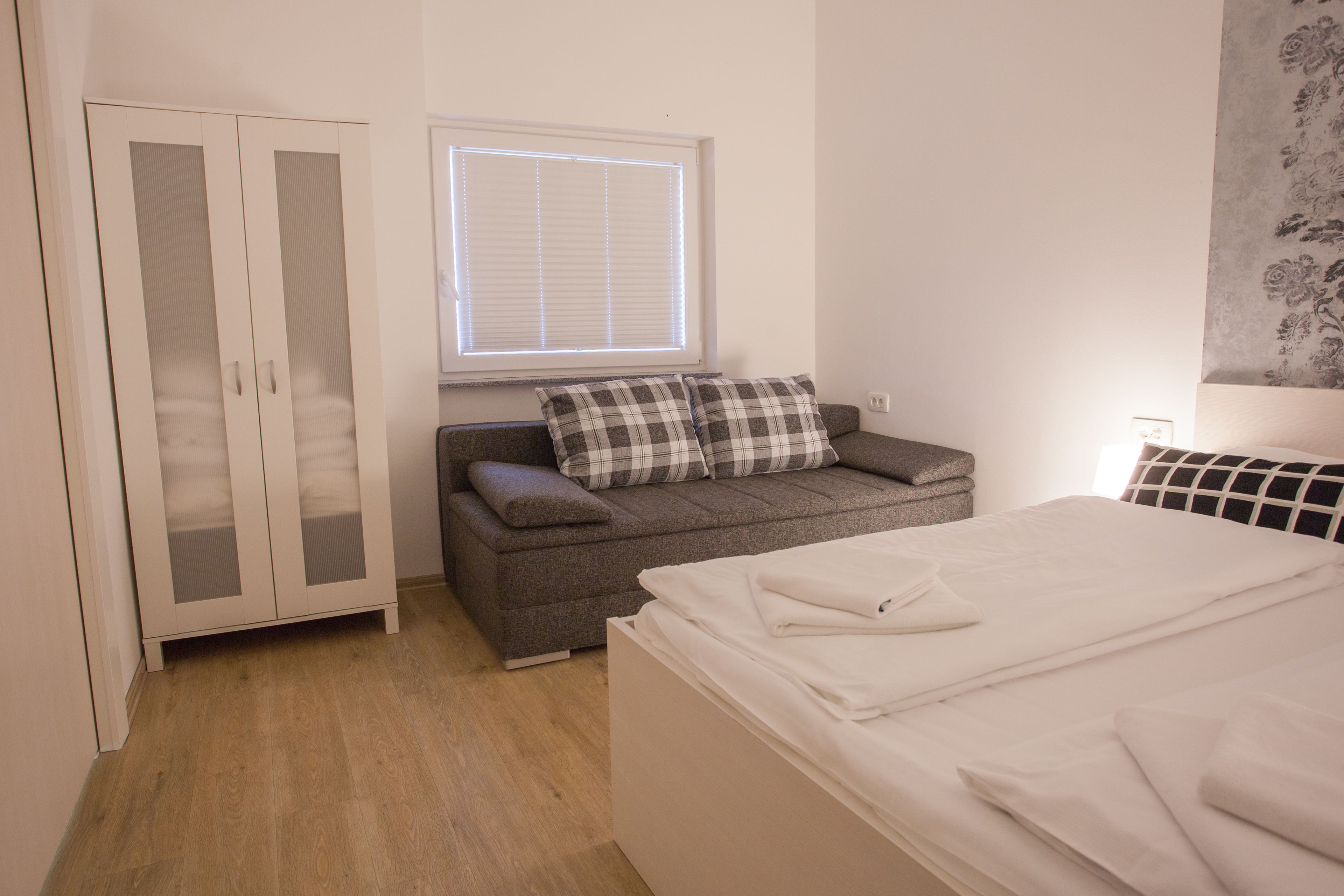 A bedroom with a wardrobe, a sofa, a bed, and a window with blinds.