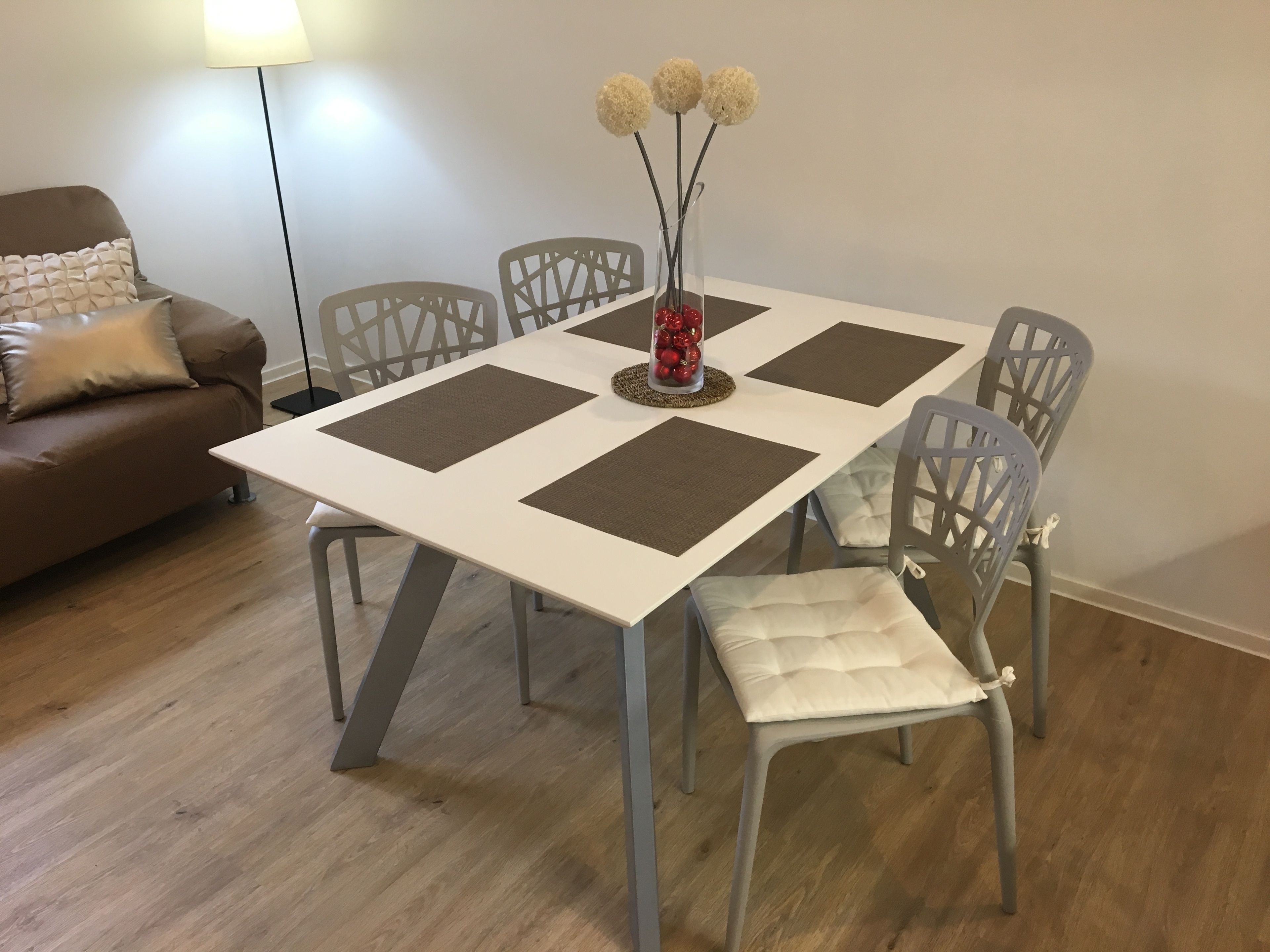 Mesa de jantar com quatro jogos americanos, rodeada por seis cadeiras. Um vaso com flores e um objeto decorativo estão sobre a mesa.