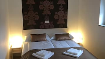 Et hotelværelse med en stor seng, to sengeborde med lamper, en mørk blomstret sengegavl og et vindue til højre.