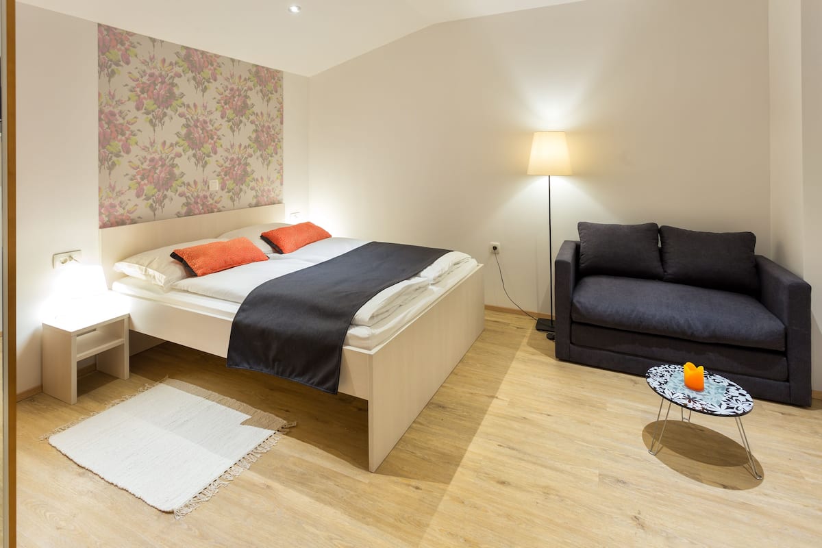 Een slaapkamer met een bed, nachtkastje, lamp, bank en een klein tafeltje met een decoratief object.