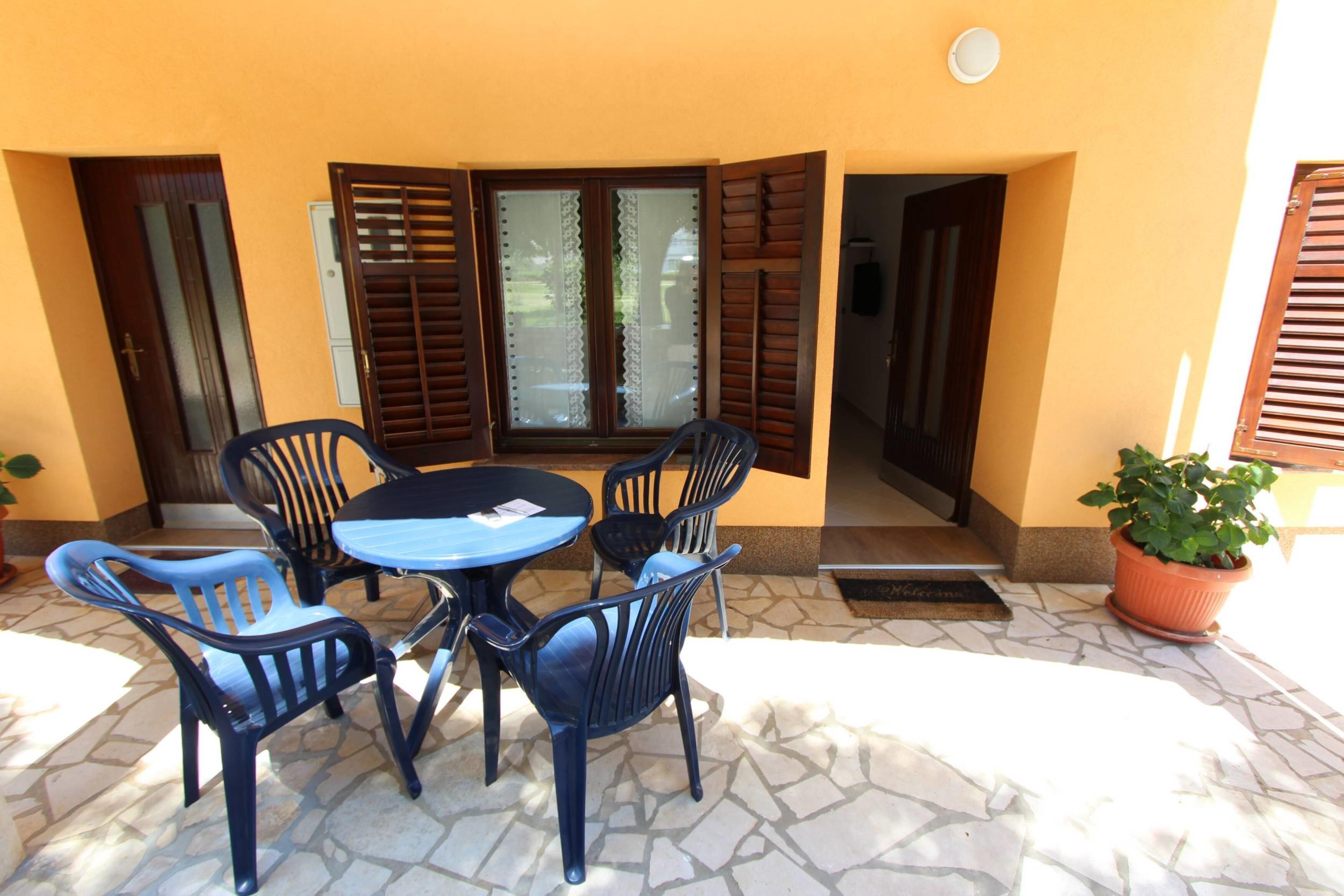 Un área de estar al aire libre con una mesa, sillas, una planta en maceta y una puerta con contraventanas abiertas.
