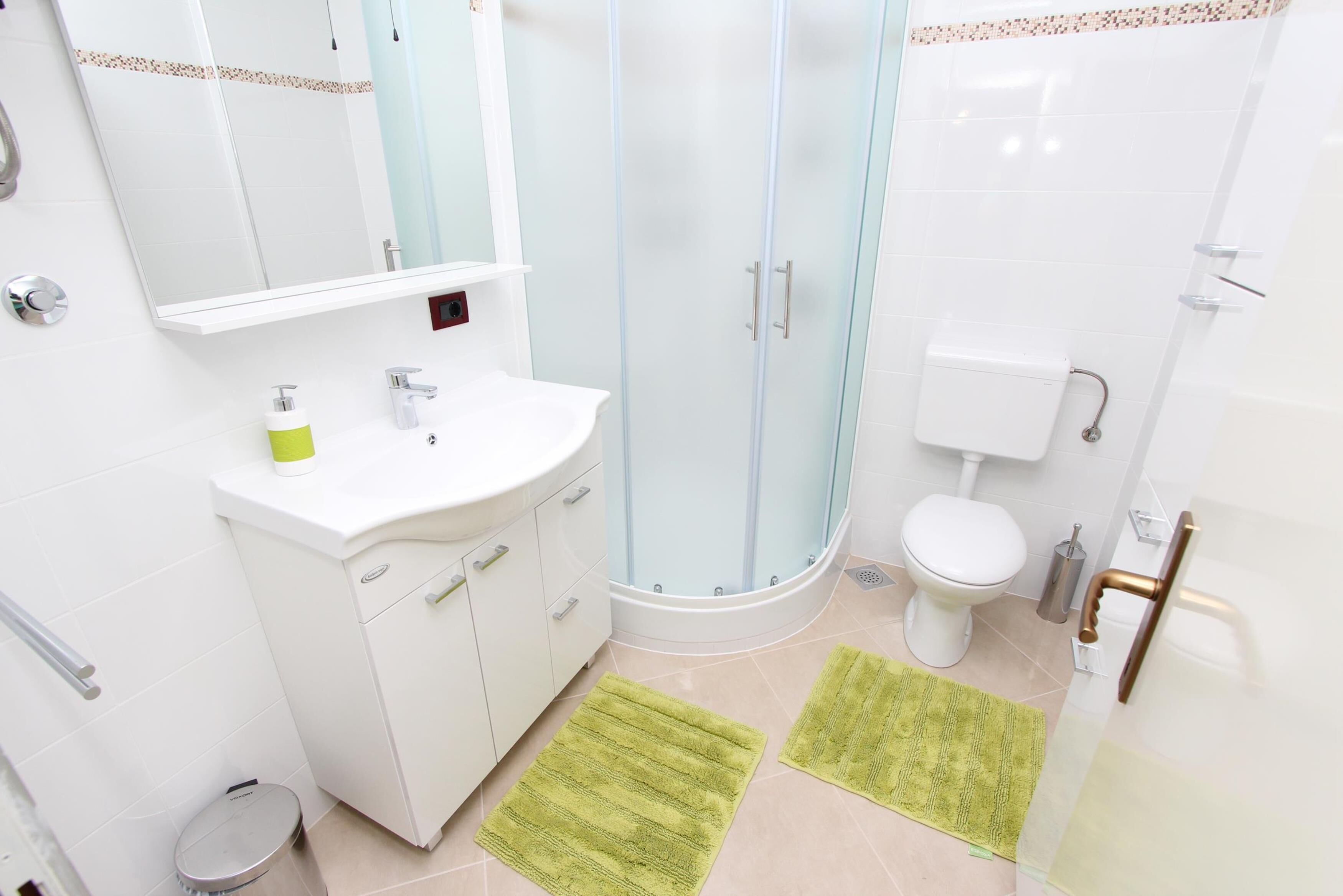 Une salle de bain moderne avec un lavabo blanc, un miroir et des toilettes, une douche en verre et deux tapis verts sur le sol.