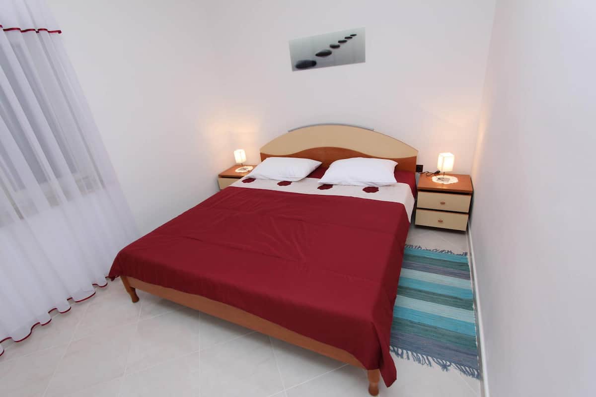 Un lit bien fait, recouvert d’une couverture rouge, avec deux oreillers blancs et une tête de lit en bois. On y trouve deux tables de chevet avec des lampes, et un tableau représentant des pierres, accroché au mur.