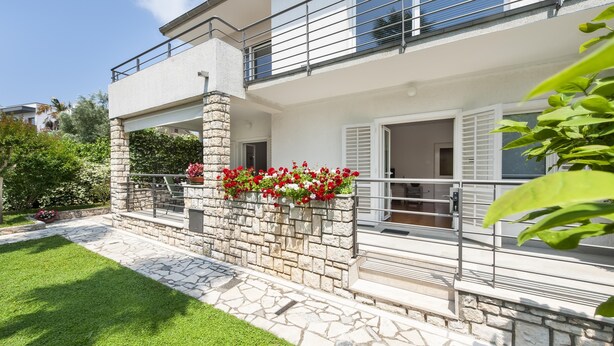 Una casa de dos pisos con fachada de piedra, un balcón con barandillas de metal y un jardín con flores rojas.