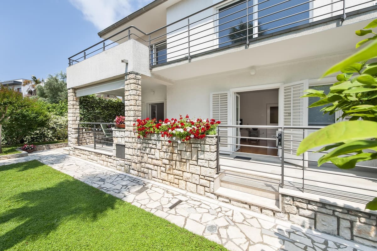 Uma casa de dois andares com fachada de pedra, uma varanda com grades de metal e um jardim com flores vermelhas.