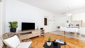 Una sala de estar moderna con una televisión de pantalla plana, una silla de mimbre, plantas en macetas y un comedor con una mesa y sillas blancas.
