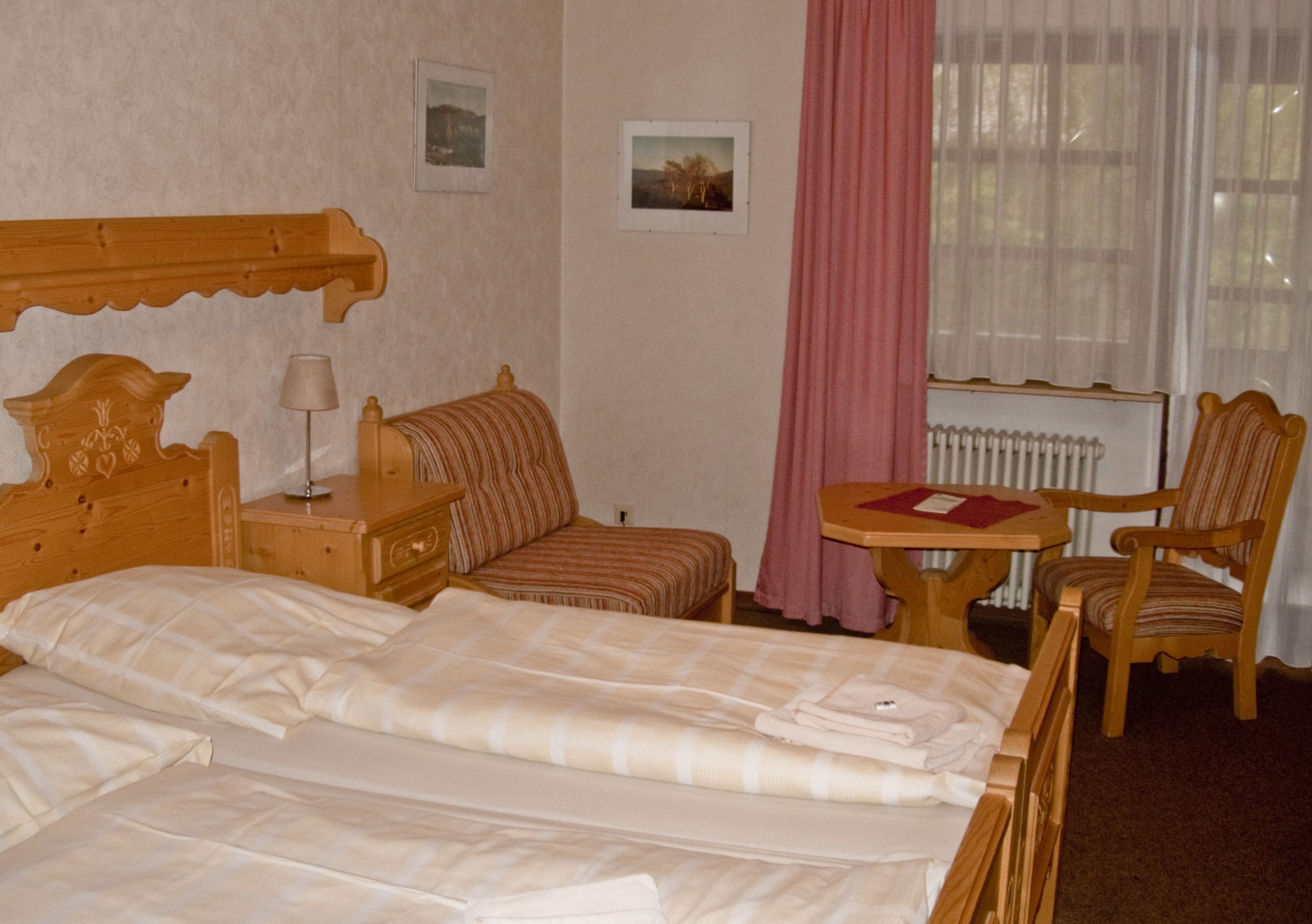 A hotel room with a large bed, a wooden headboard, a bedside table with a lamp, a striped chair, a small table with a red folder, and a window with sheer curtains.