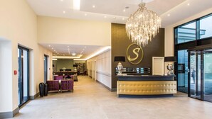 A hotel lobby with a reception desk, a chandelier, a large window, and a seating area.
