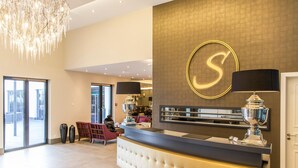A hotel reception area with a large, illuminated letter 'S' on the wall, a reception desk, and a chandelier.