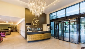 A hotel reception area with a large chandelier, a reception desk, a glass door entrance, and a seating area with a purple chair.