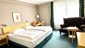 Une chambre d’hôtel avec un lit, une chaise, une petite table, une table de chevet, une lampe, un tableau encadré et une fenêtre avec des rideaux.