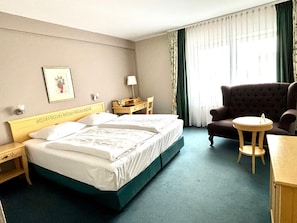 A hotel room with a bed, a chair, a small table, a nightstand, a lamp, a framed picture, and a window with curtains.