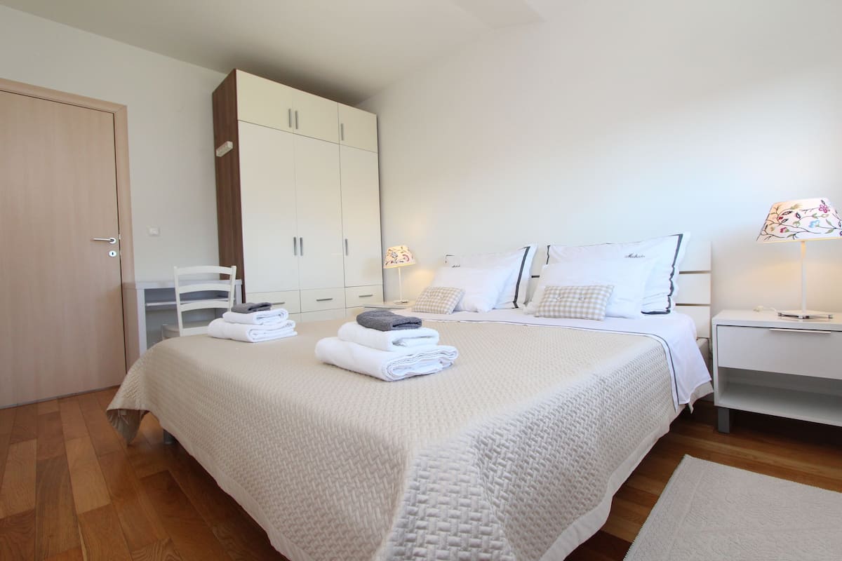 Una cama bien hecha con colcha blanca, un armario de madera y una mesita de noche con una lámpara.