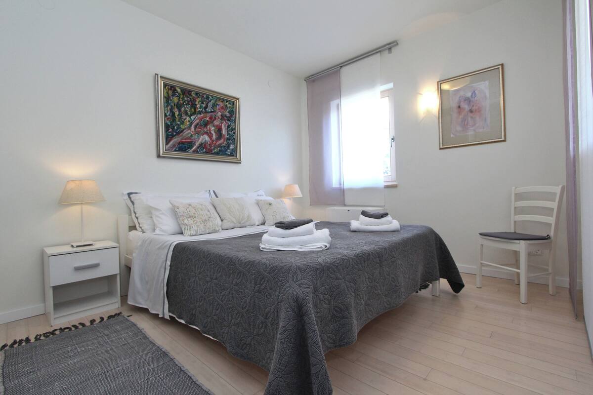 Un dormitorio con una cama, dos mesitas de noche, una silla y obras de arte en las paredes.