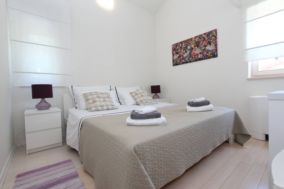 Un dormitorio moderno con una cama, dos mesitas de noche, una ventana y un cuadro abstracto y colorido en la pared.