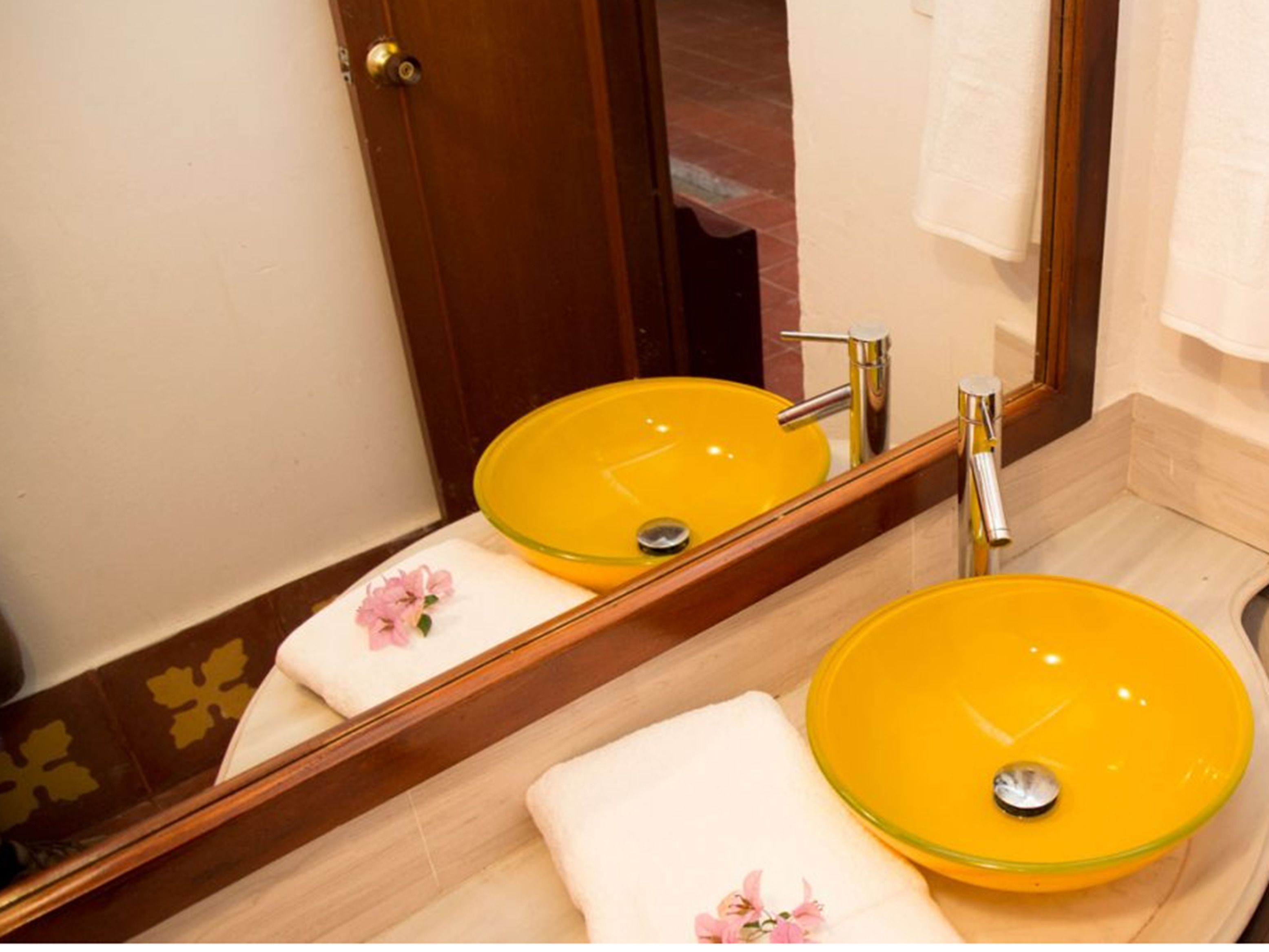 Un baño con un lavabo de vidrio amarillo, una puerta de madera y un espejo que refleja el lavabo y una toalla con un estampado de flor rosa.