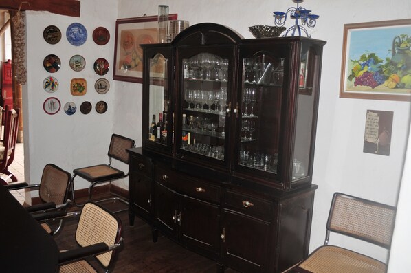 Een kamer met een donker houten kast vol glazen en flessen, een wand versierd met diverse borden, en rieten stoelen.