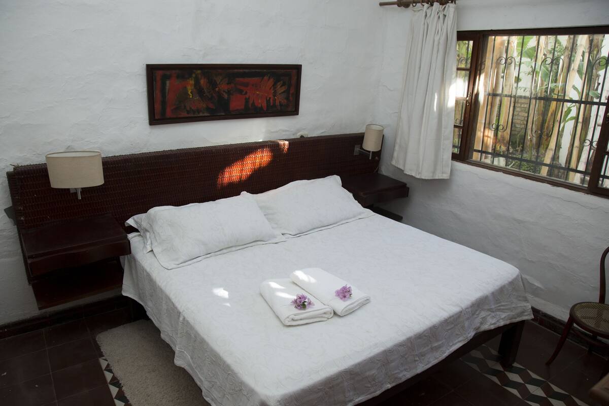 Una cama bien hecha con ropa blanca y toallas, una obra de arte enmarcada en la pared, una ventana con vistas a la vegetación y una silla en la esquina.