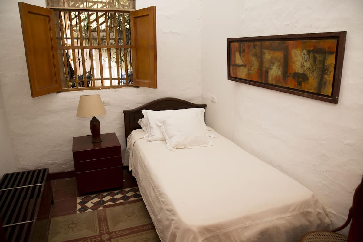 Un dormitorio pequeño y ordenado con una cama individual, una mesita de noche con una lámpara, una ventana con contraventanas abiertas y una obra enmarcada en la pared.