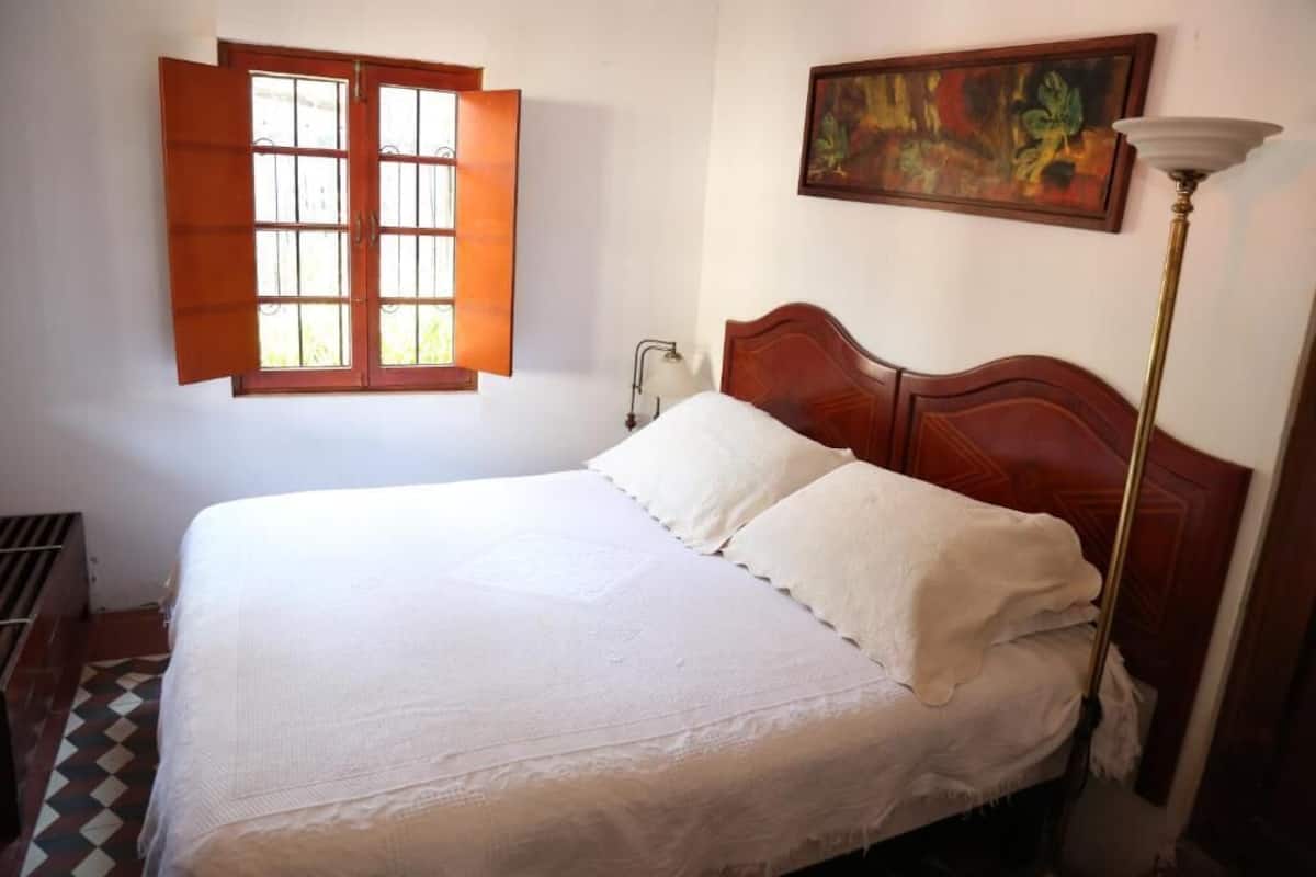Una cama bien hecha con sábanas blancas, cabecera de madera, ventana con contraventanas abiertas, una lámpara de pie y un cuadro en la pared.
