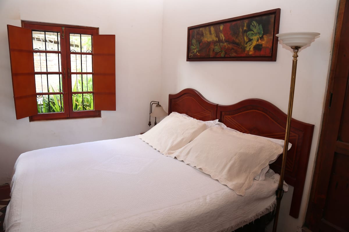 Un dormitorio con cama de madera, ropa de cama blanca, una ventana con contraventanas abiertas, una lámpara de noche y un cuadro en la pared.