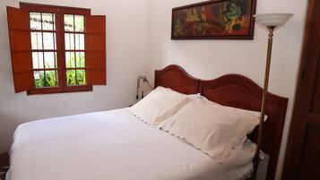Quarto com cama de madeira, roupa de cama branca, janela com portadas abertas, abajur e um quadro na parede.