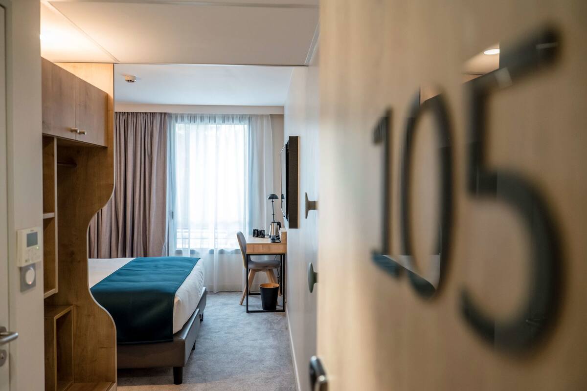 A hotel room with a bed, a desk, a chair, a window with curtains, and a door with the number 105.
