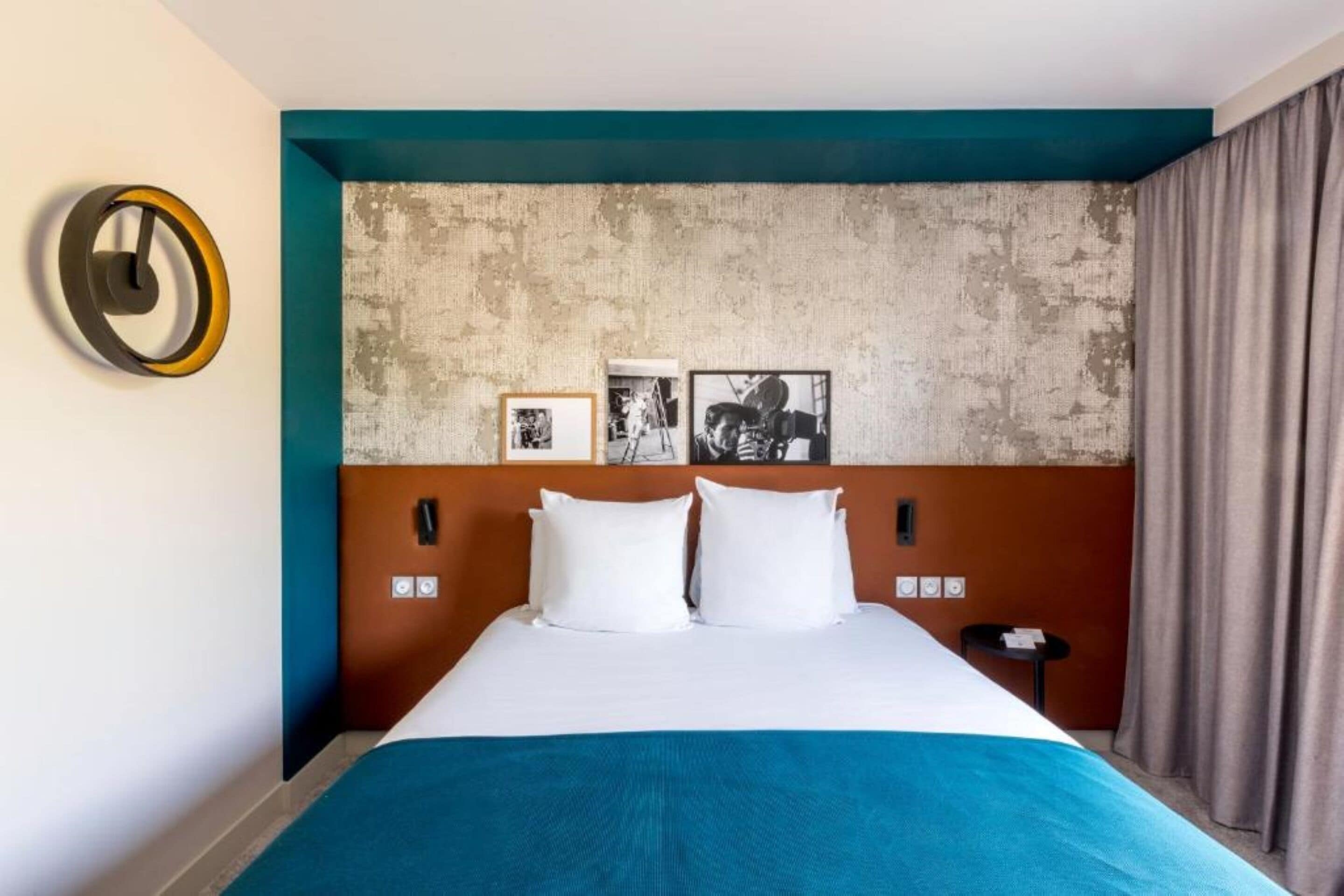 A hotel room with a large bed, a headboard with artwork, a clock, and a small table with a chair.