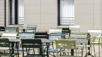 An outdoor seating area with tables and chairs, a modern building with large windows, and a tree overhead.