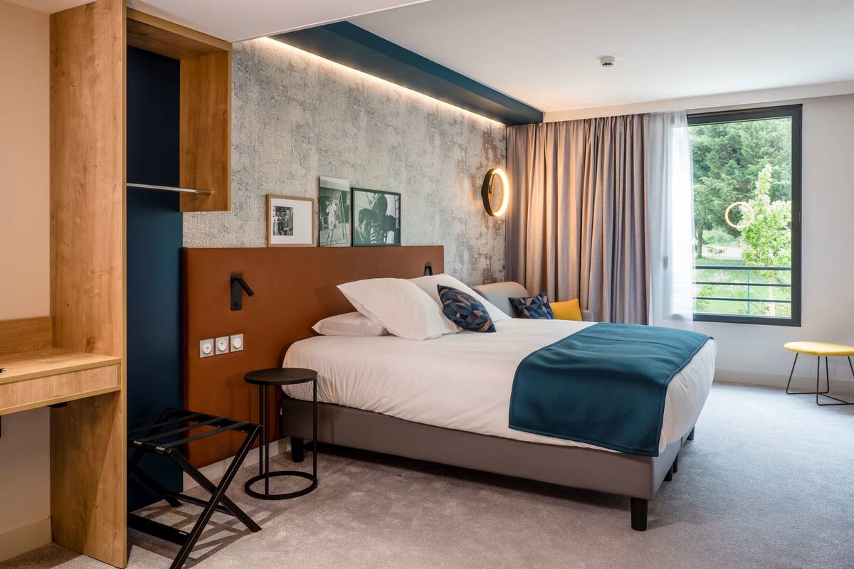 A modern bedroom with a bed, a desk, and a window with a view of greenery.