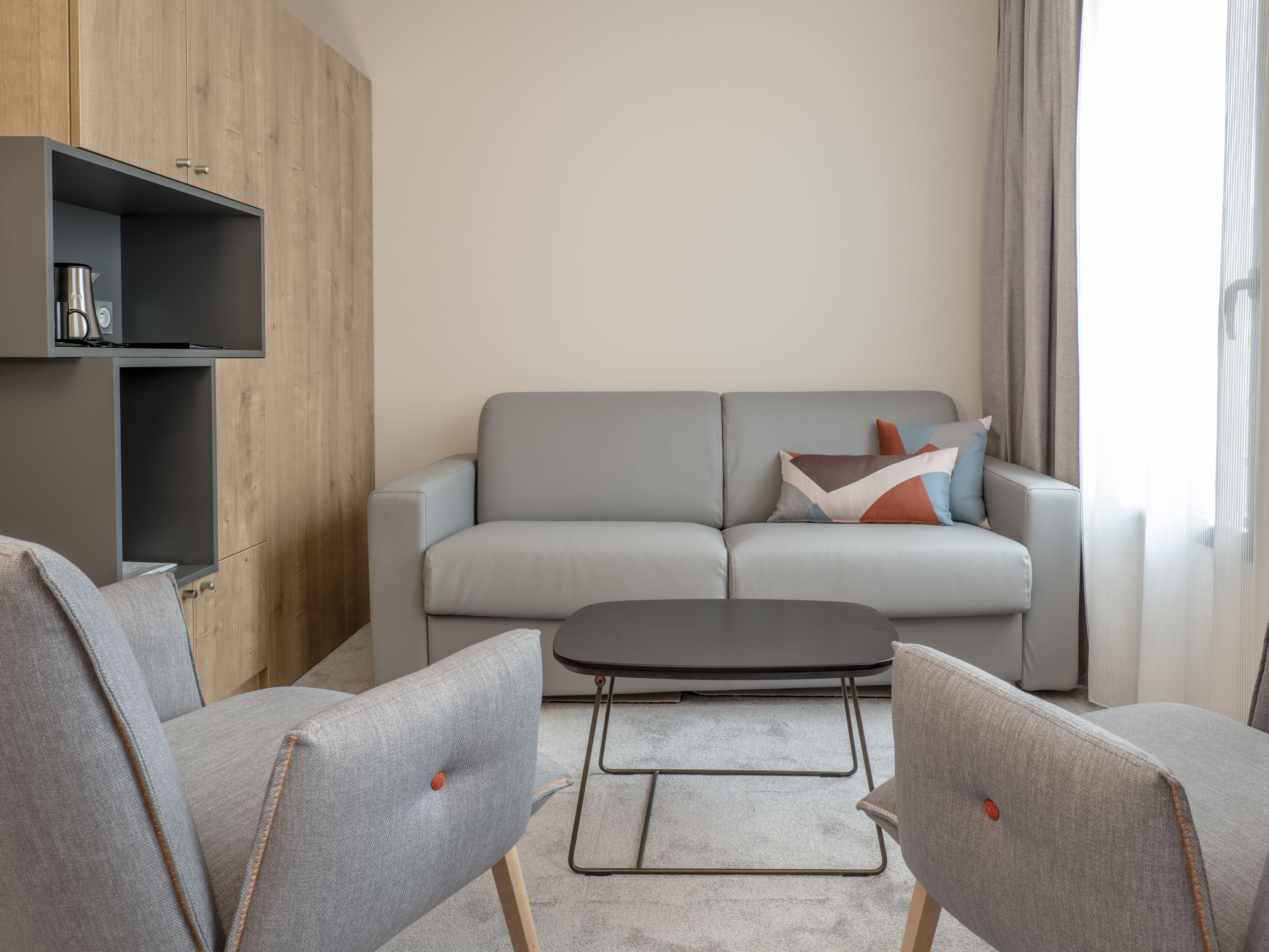 A modern living room with a grey sofa, two grey armchairs, a small coffee table, and a wooden cabinet with a coffee machine.